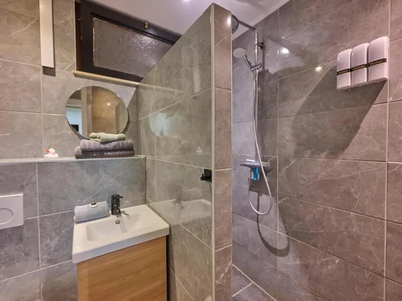 Badkamer met grijze tegels, wastafelmeubel met houten front en een inloopdouche met regendouche.