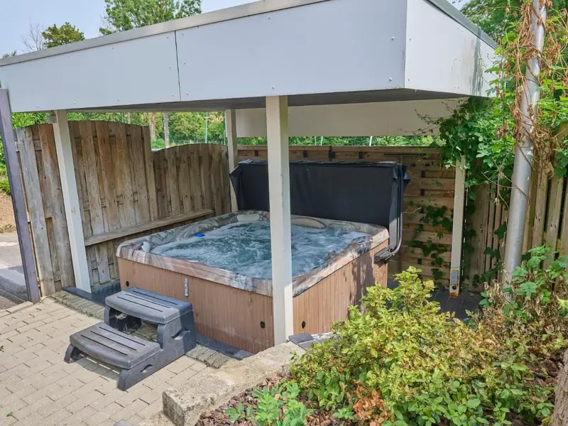 Jacuzzi onder een wit afdak met houten schuttingen, een opstapje en struiken op de voorgrond.