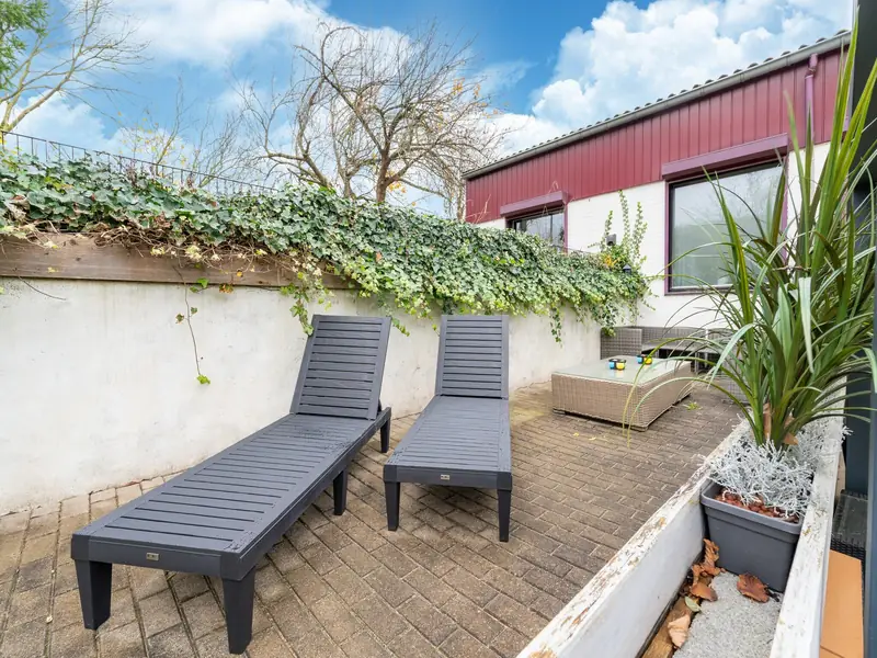 Paved patio with two grey sun loungers, a white wall with ivy and blue sky.
