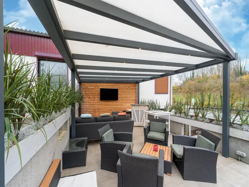 Covered veranda with wicker lounge chairs, a lounge sofa and a TV, trees and grass beyond.