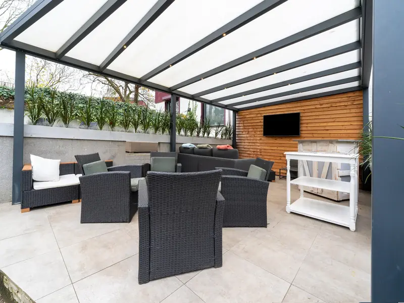 Covered veranda with dark wicker lounge chairs, a lounge sofa and a TV on a wooden wall.
