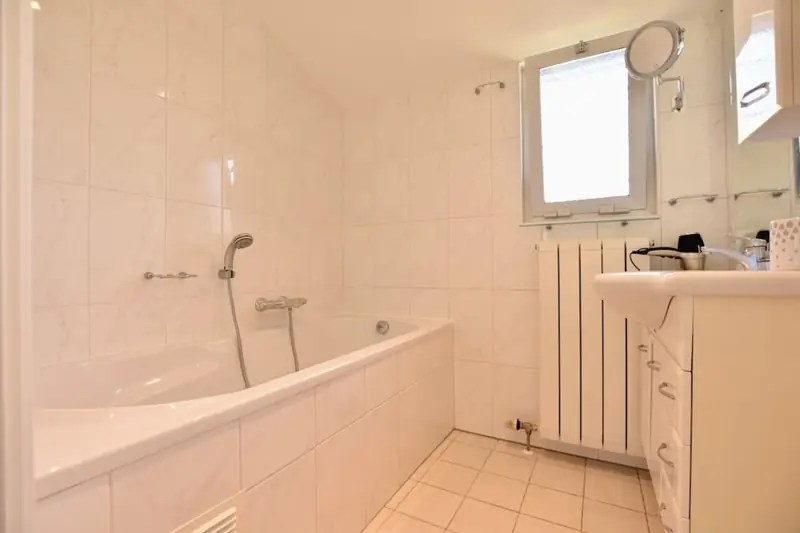 Weiß gefliestes Bad mit Badewanne und Handbrause, Heizkörper, Waschtisch und kleinem Fenster.