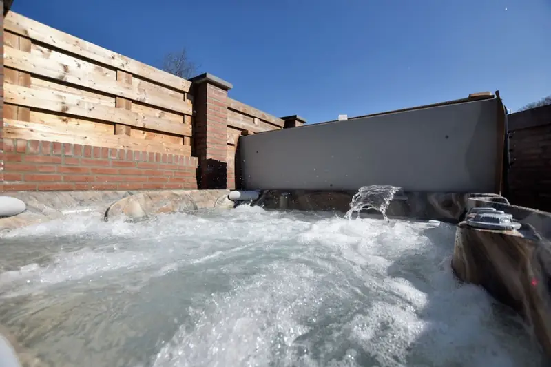 Bruisend bubbelbad met waterstraal, hoofdsteunen en opgeklapt deksel onder een blauwe lucht.