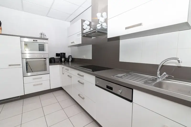 Küche mit weißen Schränken, Kochfeld, Spüle, Geschirrspüler, Einbaubackofen und Kaffeemaschine.
