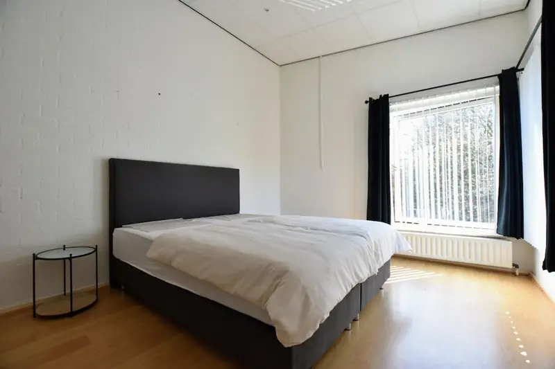 Schlafzimmer mit Doppelbett, rundem Beistelltisch und großem Fenster mit Lamellenvorhang.