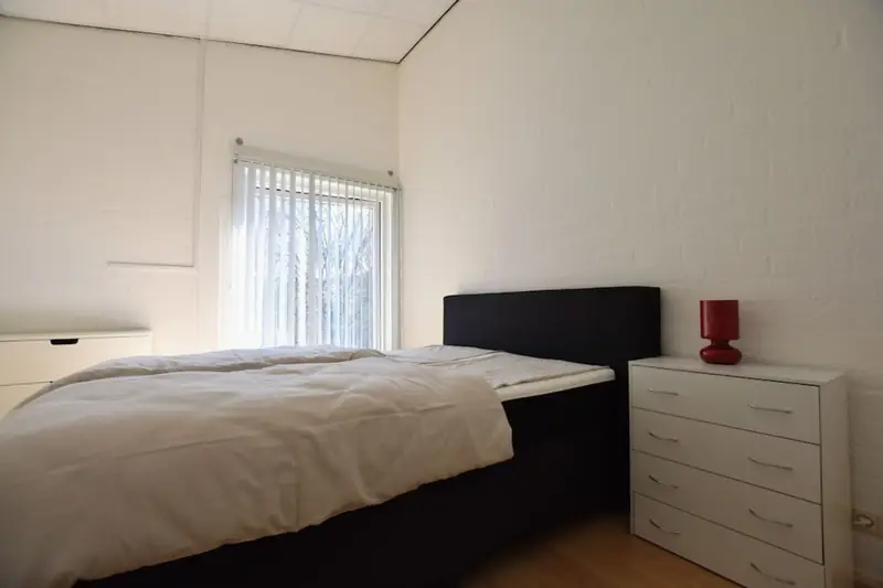 Schlafzimmer mit Doppelbett, weißer Kommode mit roter Lampe und Fenster mit Lamellenvorhang.
