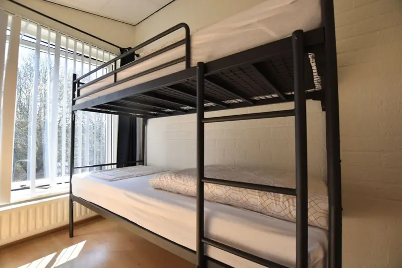 Stapelbed met zwart metalen frame en ladder naast een raam met verticale lamellen.