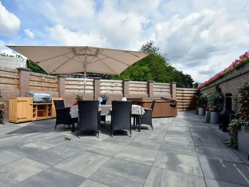 Große geflieste Terrasse mit Tisch unter Sonnenschirm, abgedecktem Whirlpool und blühenden Pflanzen.