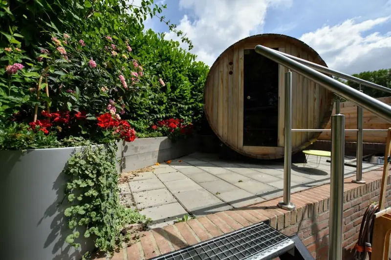 Houten tonsauna op een verhoogd betegeld terras met een stalen reling, bloemen en een haag.
