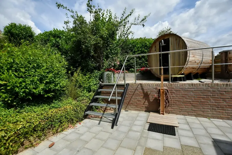 Fasssauna auf einer erhöhten Backsteinterrasse, davor eine Metalltreppe und eine Außendusche.