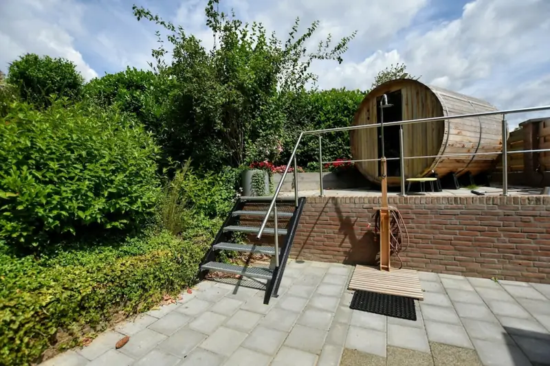 Tonsauna op een verhoogd bakstenen terras, met een metalen trap en een buitendouche ervoor.