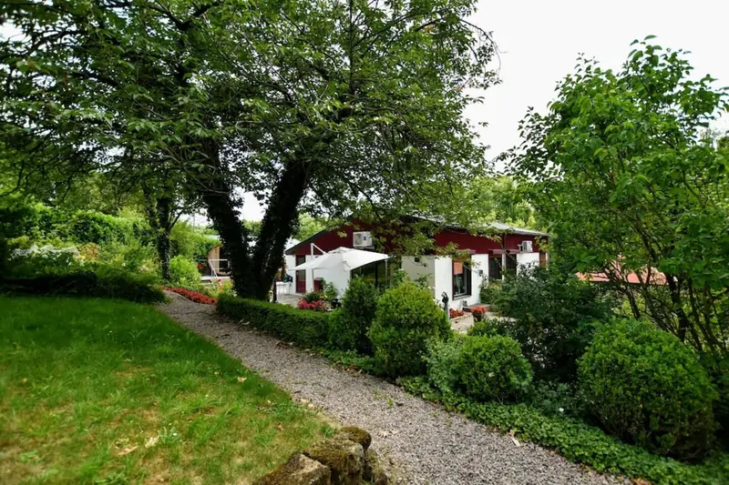 Garten mit Rasen, Kiesweg und runden Büschen; hinter den Bäumen ein weißes Gebäude mit roter Verkleidung.