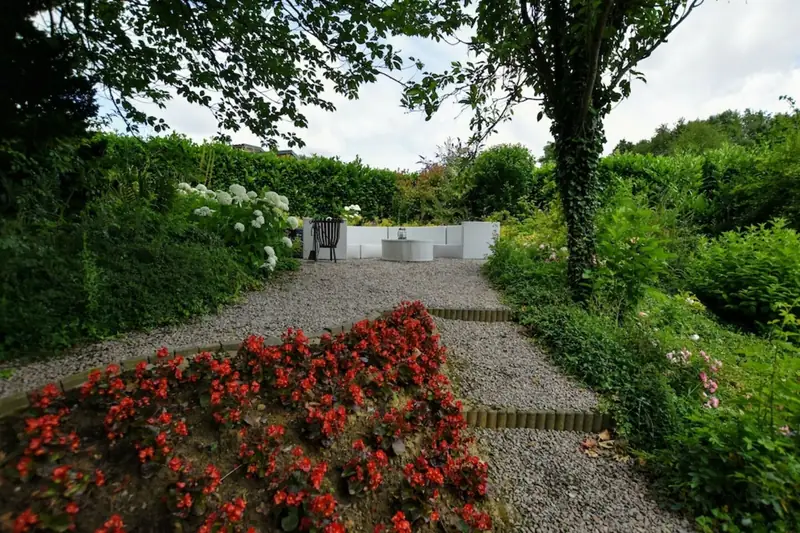 Grindpad tussen hagen leidt langs rode bloemen naar een witte zithoek met een tafel.